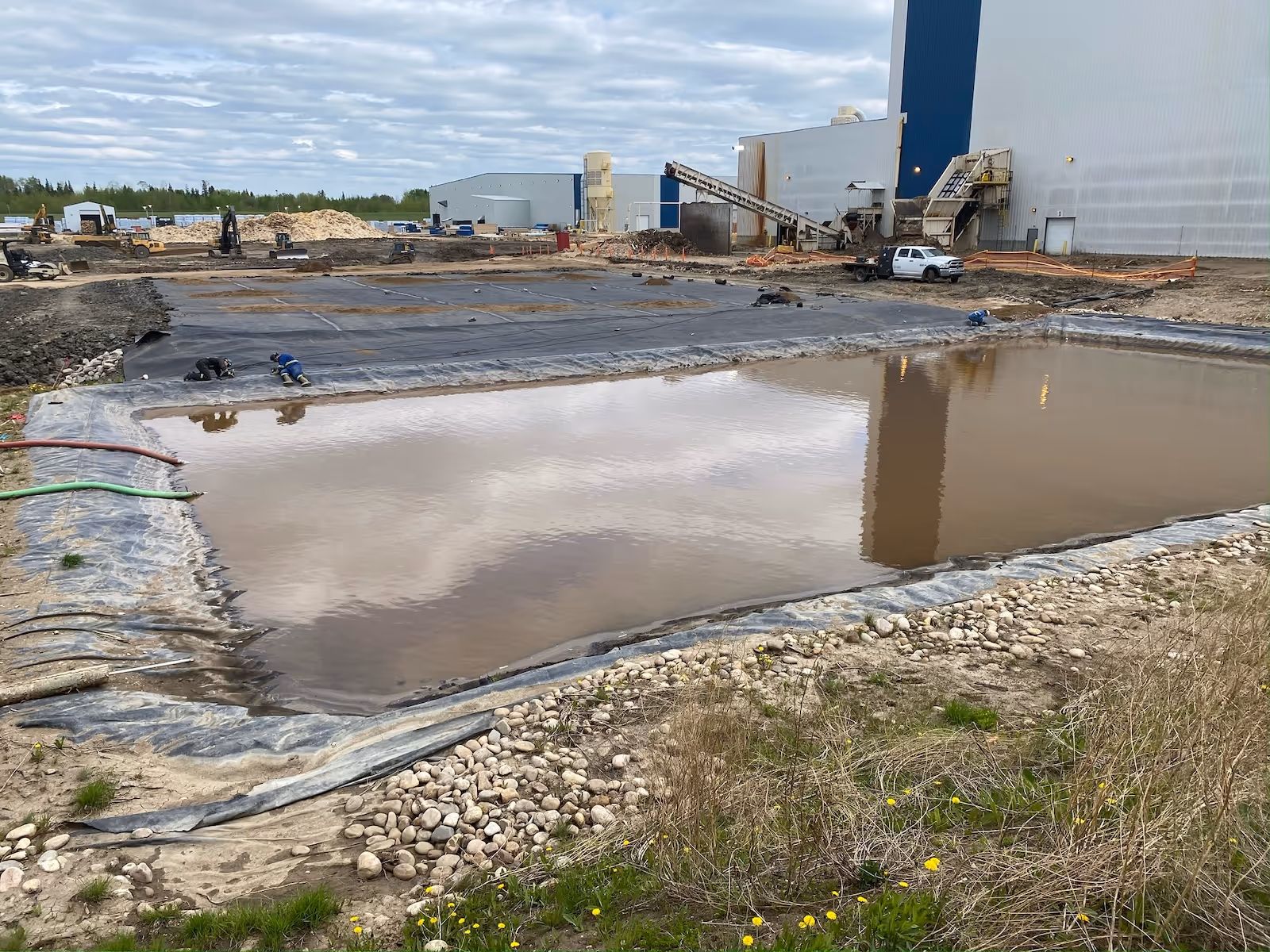 Construction site with a large water-filled pond and workers covering ground with black plastic sheets.