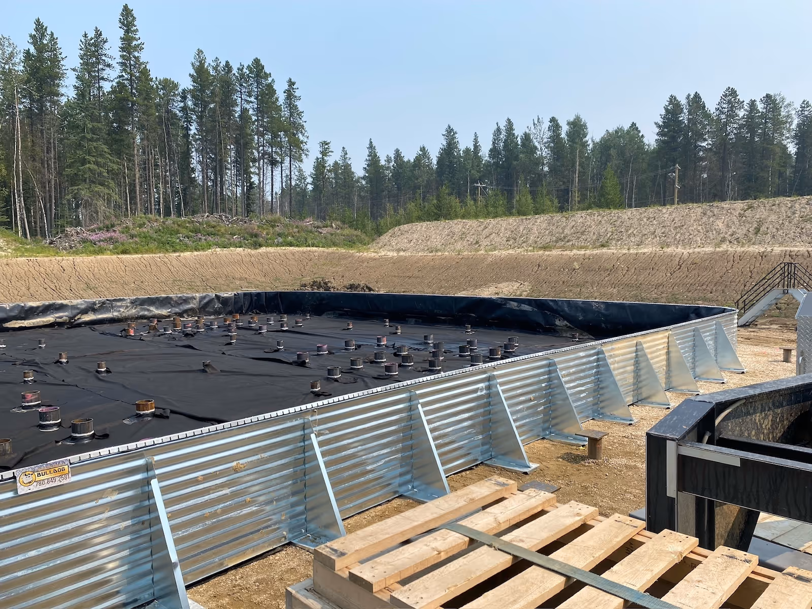 Large rectangular metal framed containment area lined with black tarp and capped pipes inside in a forest clearing.