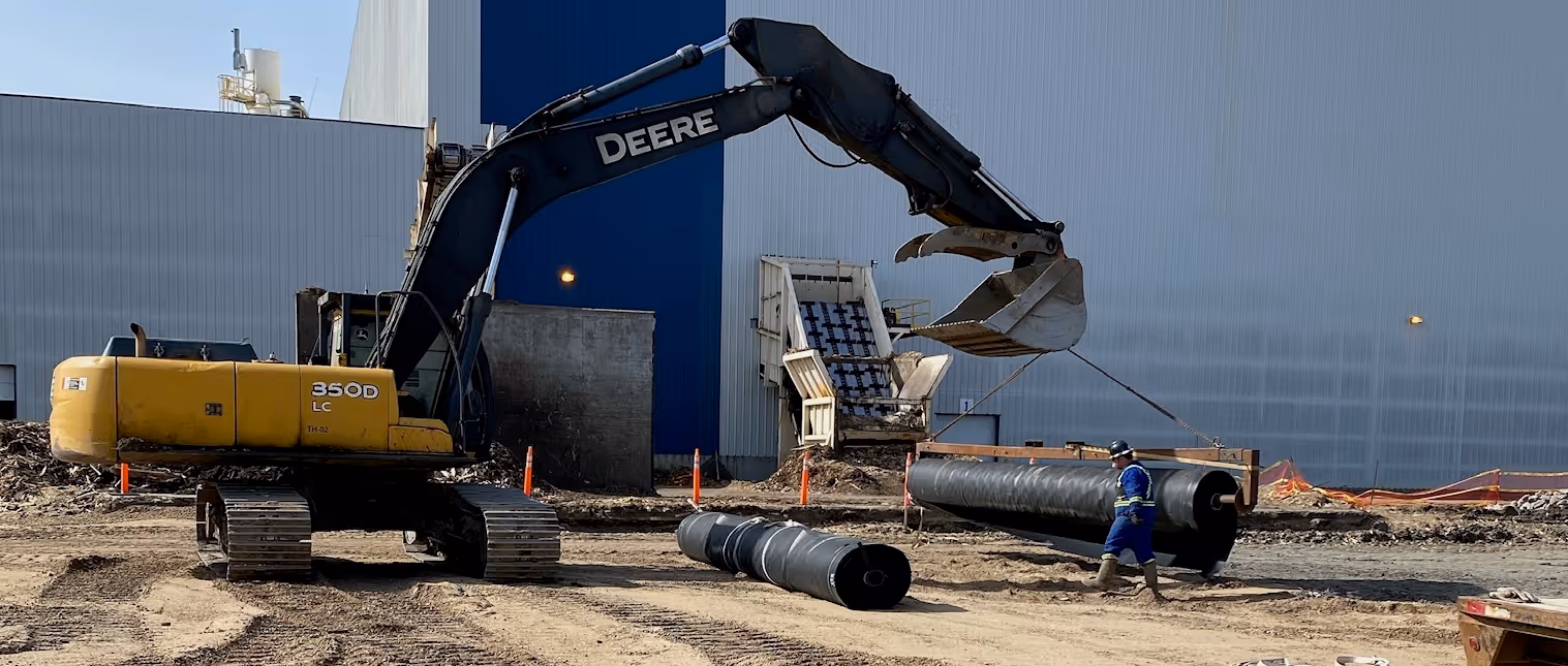 Excavator lifting a large black cylindrical pipe while a worker in safety gear walks nearby on a construction site.