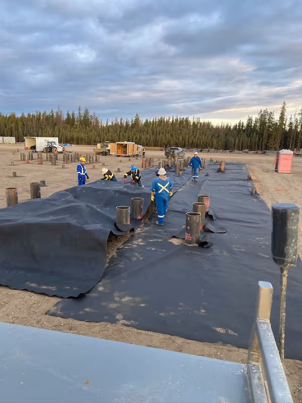 Workers in blue coveralls laying black geotextile fabric over construction ground with pipes emerging.