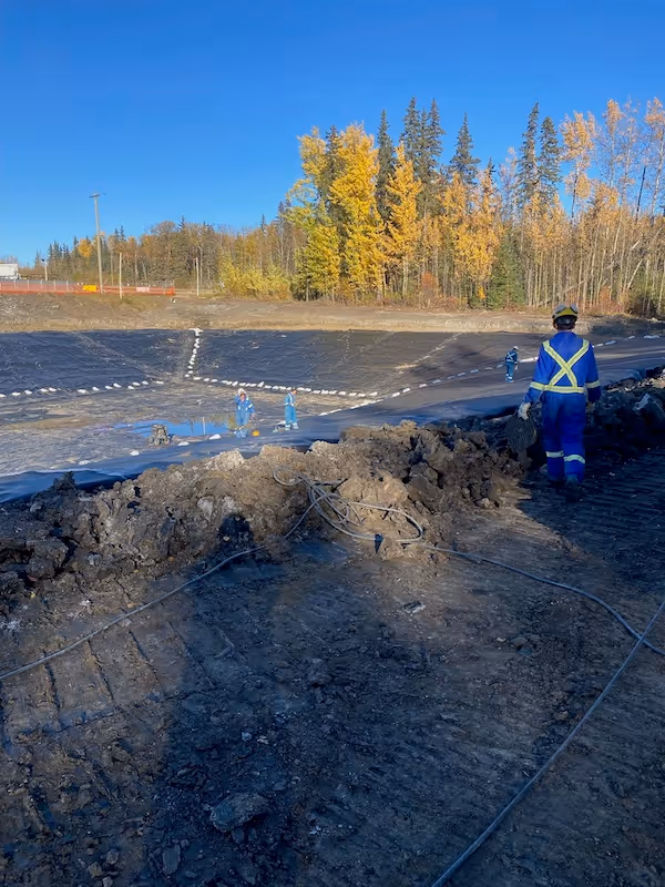 Workers in blue protective gear on a lined soil containment pond with autumn trees under a clear sky.