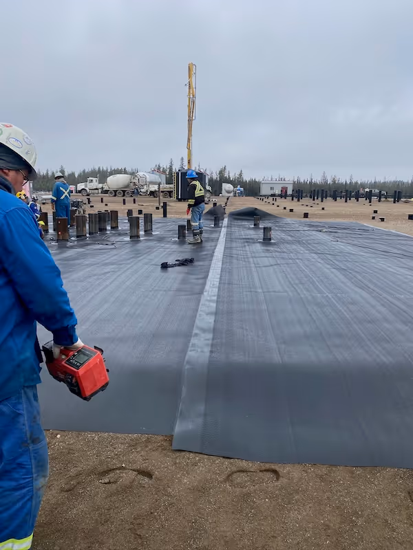 Workers installing and welding a large black liner sheet on an outdoor construction site with equipment nearby.