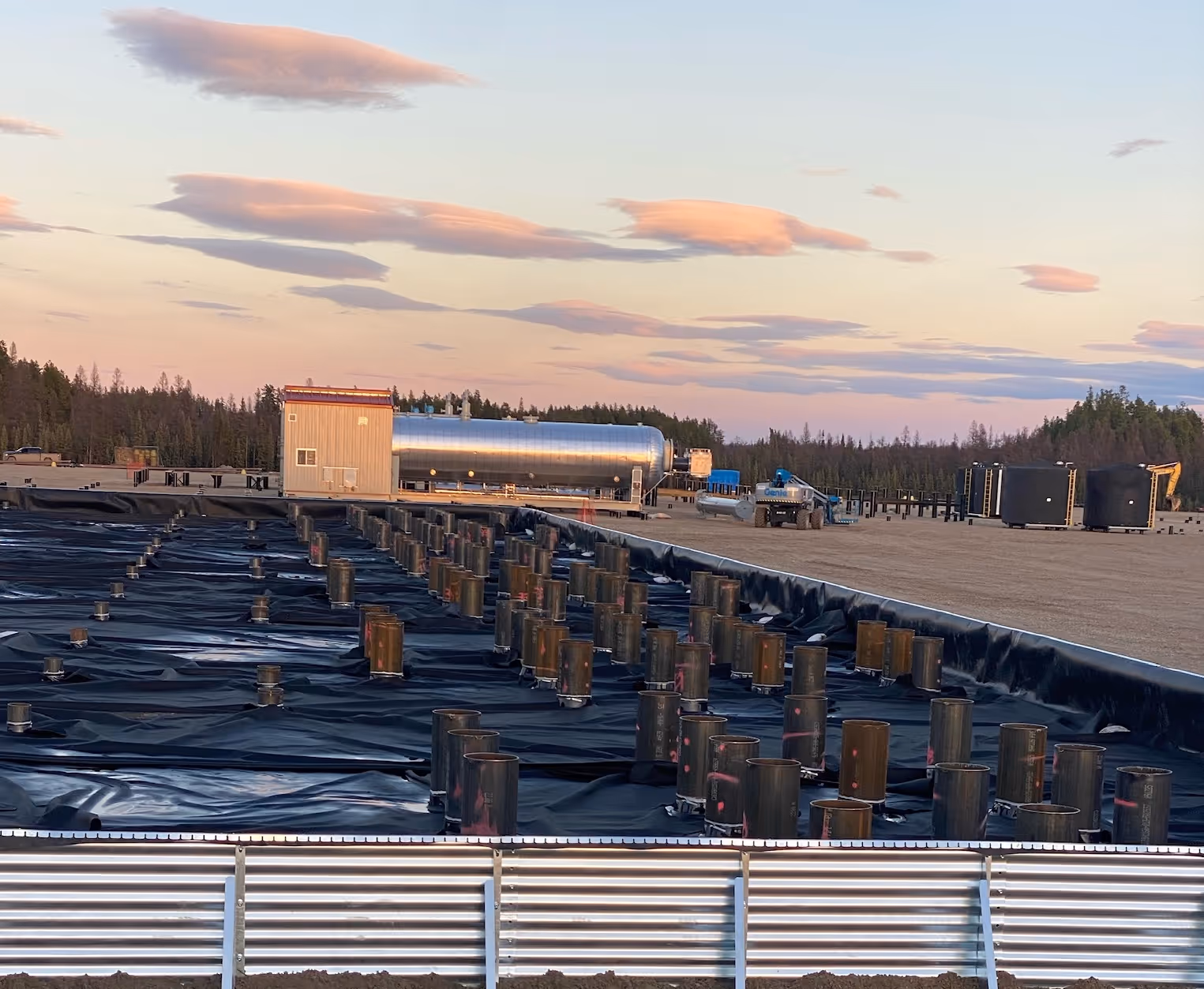 Construction site with black plastic sheeting, metal piles, and equipment at sunset near forest.