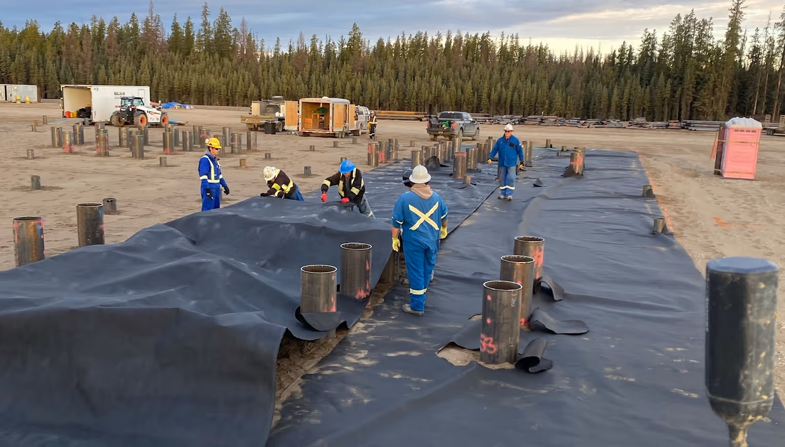 Workers in protective gear lay out black geomembrane plastic over soil with pipes protruding.