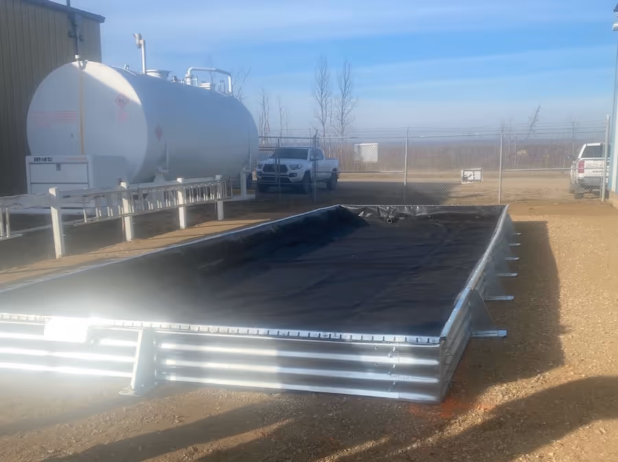 Rectangular metal containment with a black liner on gravel outside near tanks and vehicles.