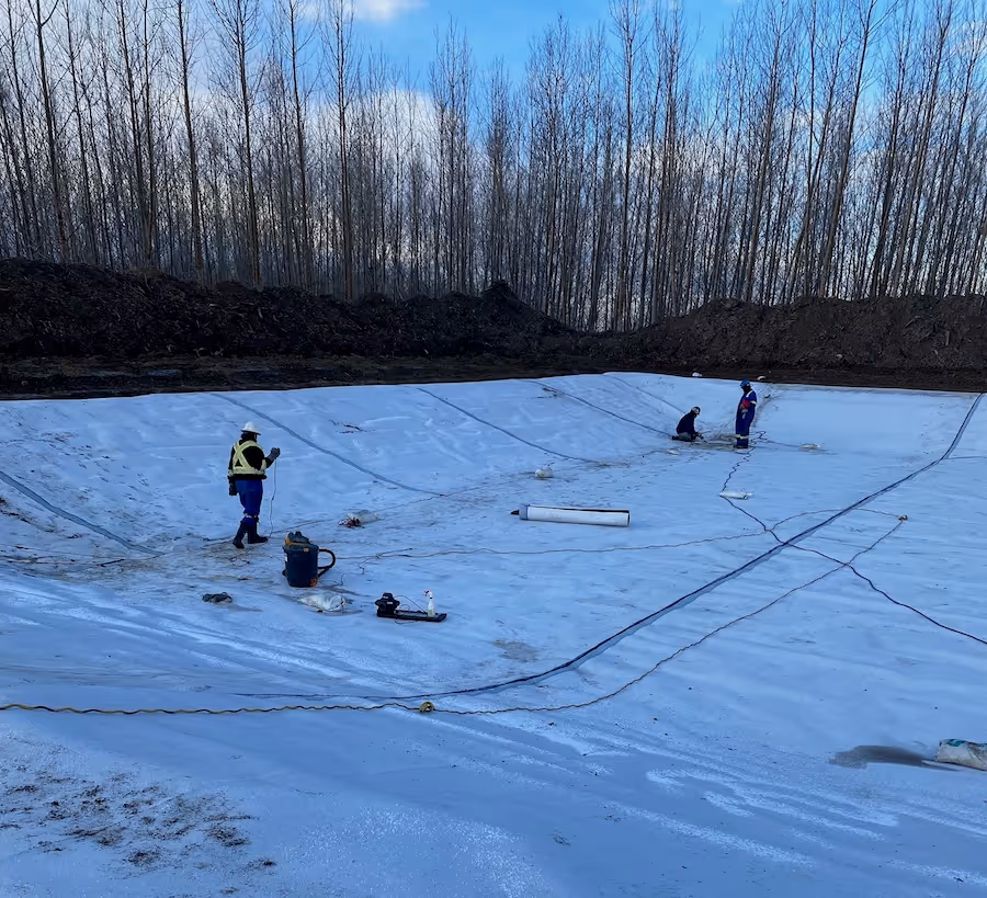 Workers installing a large white liner in an empty excavation pit surrounded by bare trees and soil.