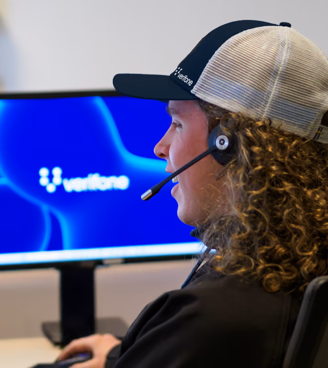 Customer support representative wearing a headset and Verifone cap working at a computer displaying the Verifone logo.