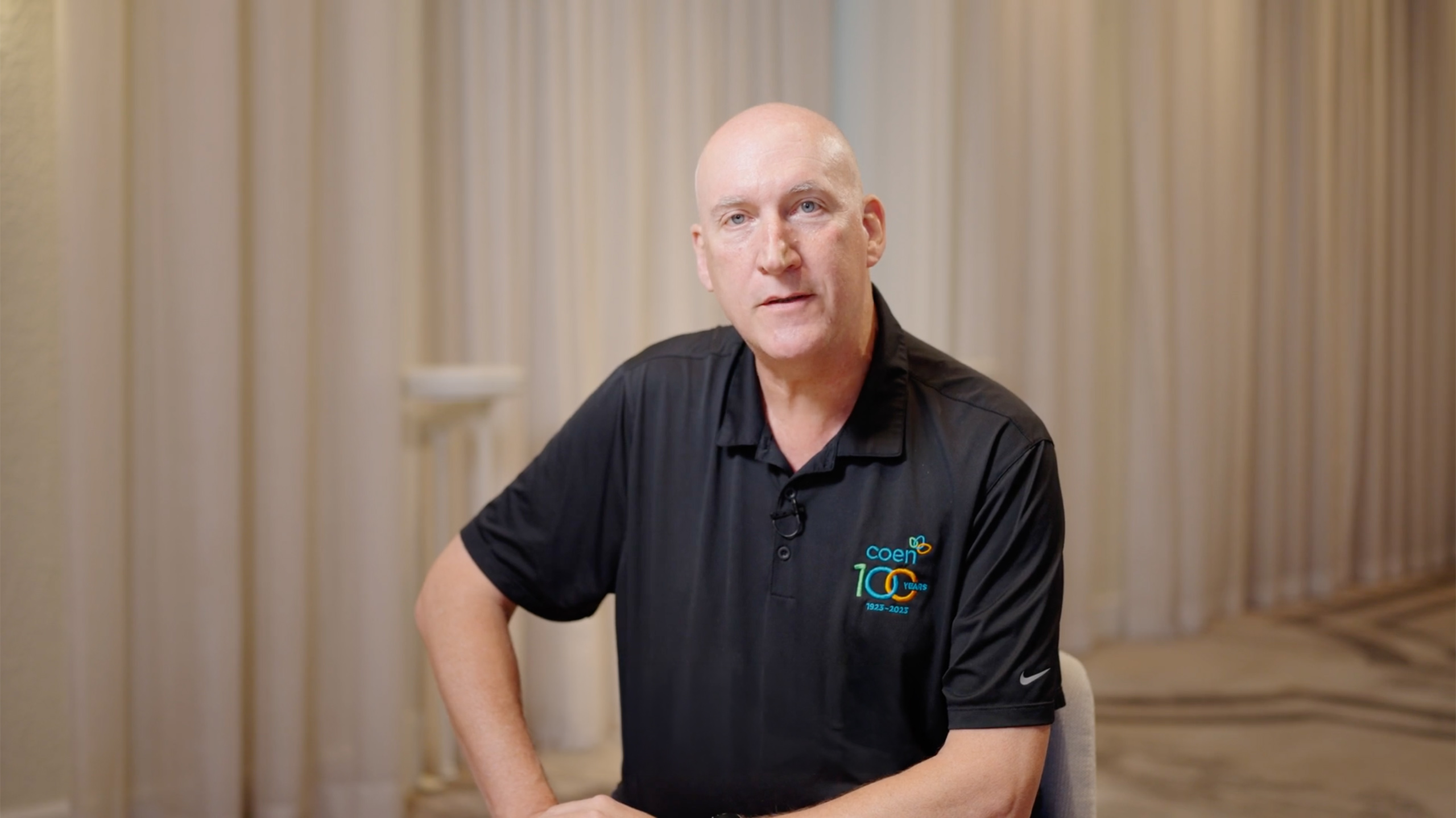 Bald man in a black polo shirt with a Coen 100 years logo speaking in an indoor setting with beige curtains.