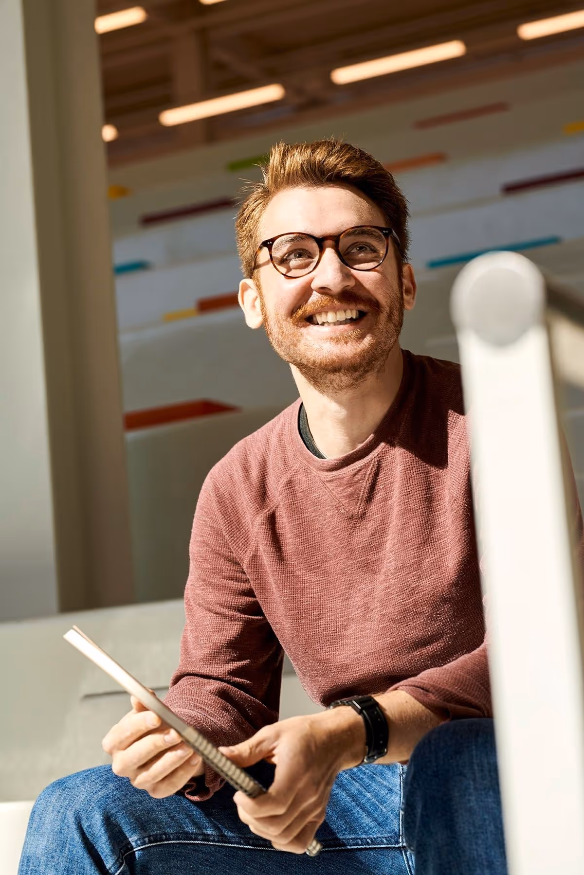 Smiling man with glasses wearing a maroon sweater and jeans, holding a notebook indoors.