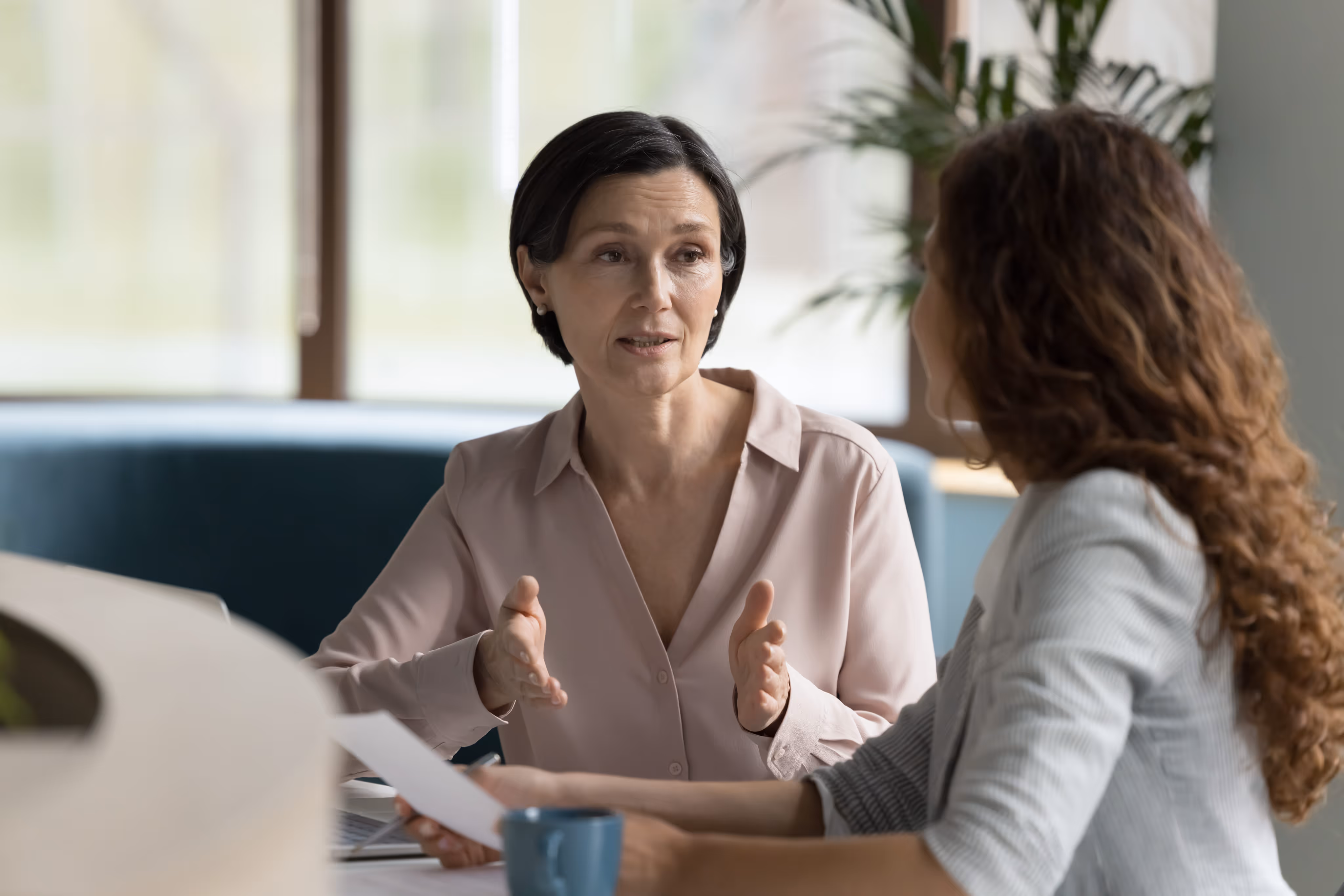 Two women having a conversation at a table in a bright office, one gesturing with hands while the other holds a paper.