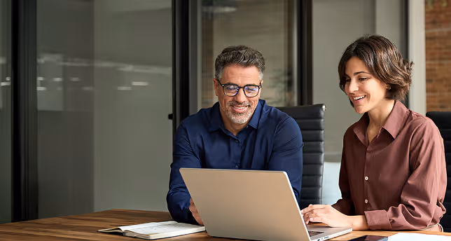 Two professionals smiling and working on a laptop at a wooden desk in a modern office.