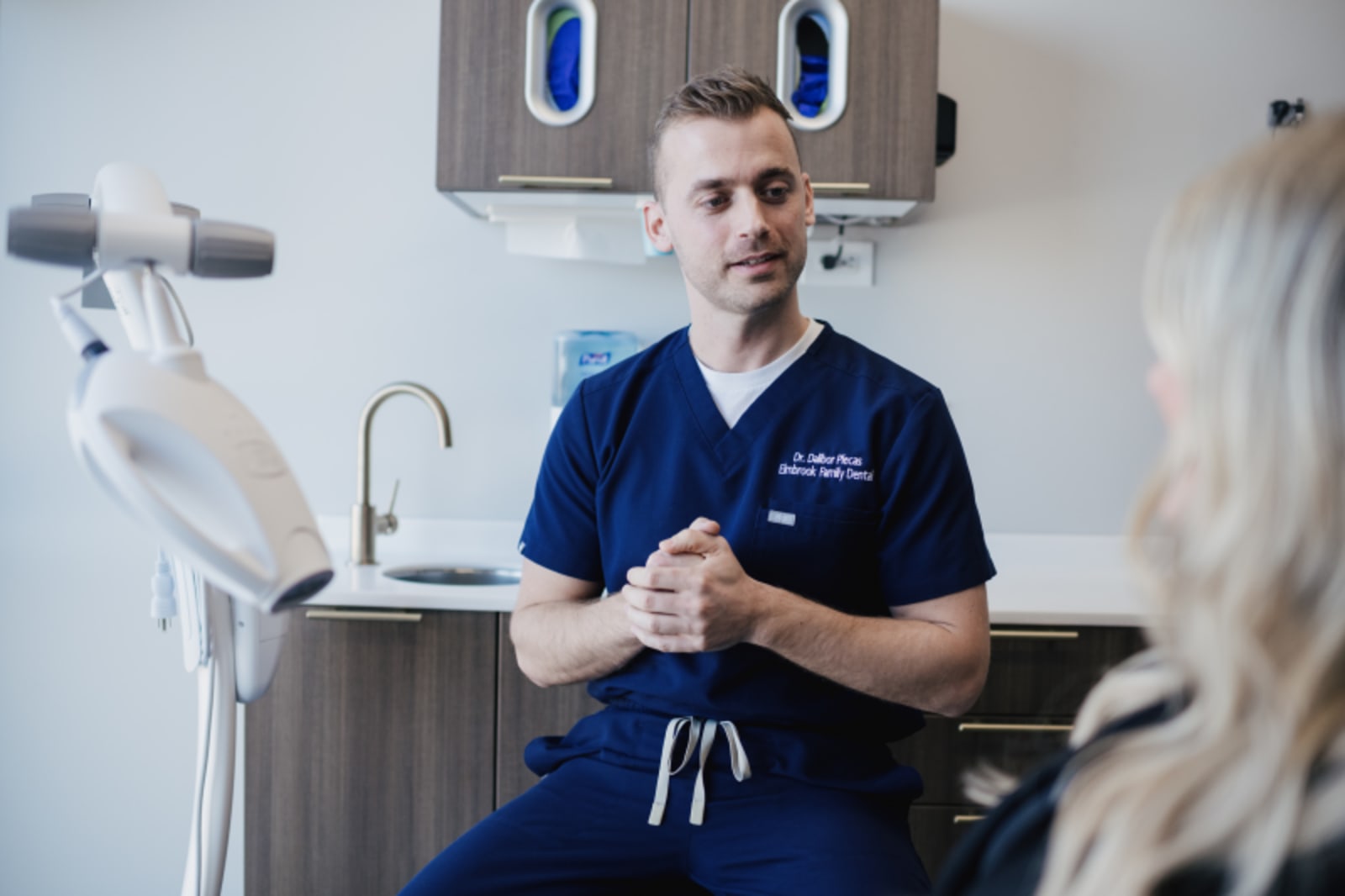 Dentist in blue scrubs holding dental model during patient consultation in treatment room.