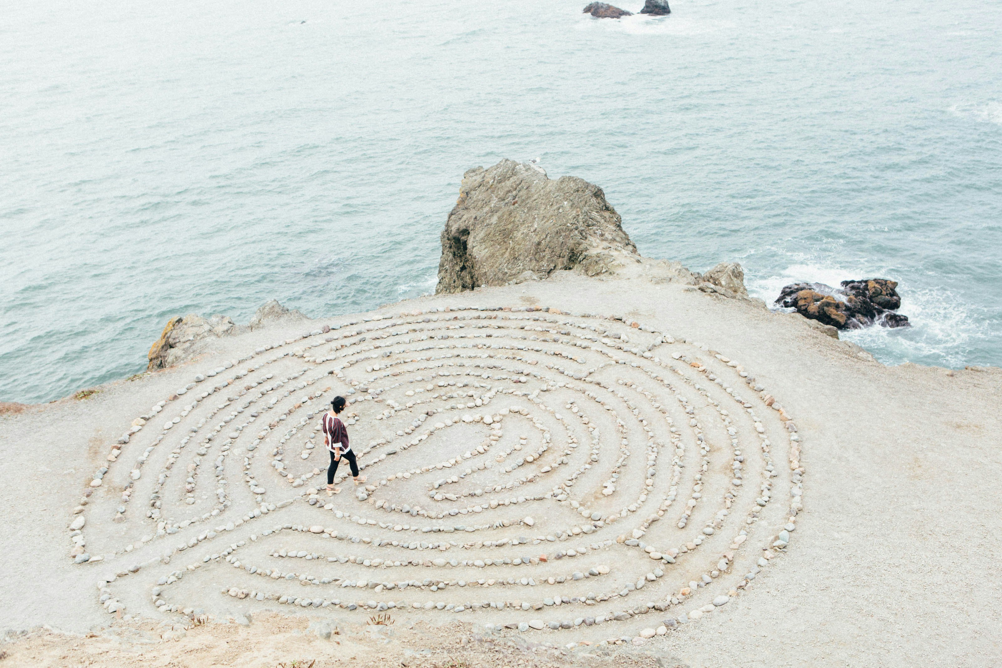 Person walking barefoot inside a stone labyrinth on a cliff overlooking the ocean.