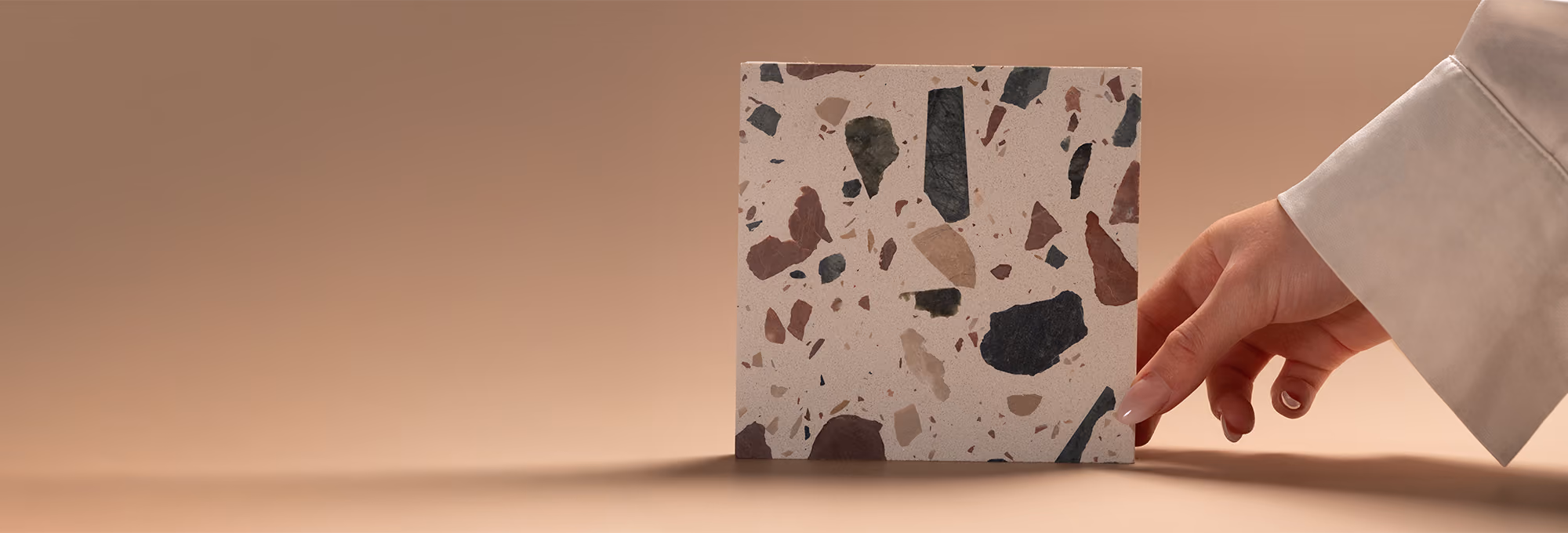 Hand holding a square terrazzo tile with various sized stone chips on a beige background.