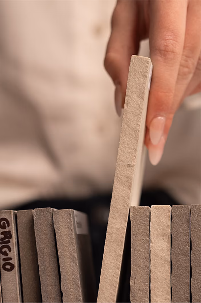 Hand with manicured nails selecting a beige tile from a vertical tile sample display.