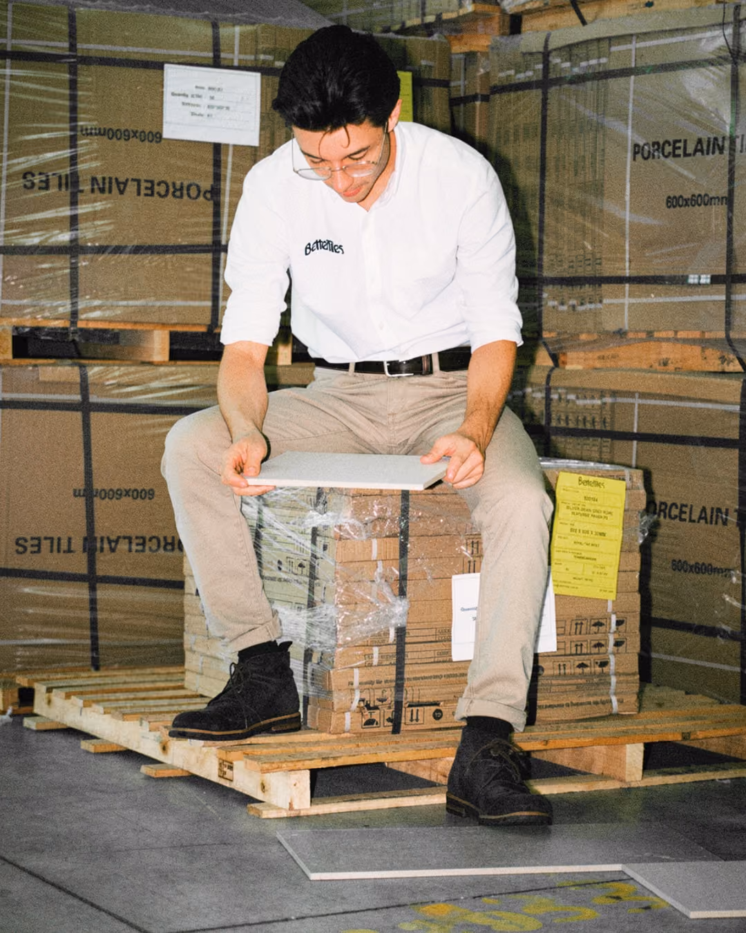 Man in glasses and white shirt sits on a pallet holding a tile sample in a warehouse.
