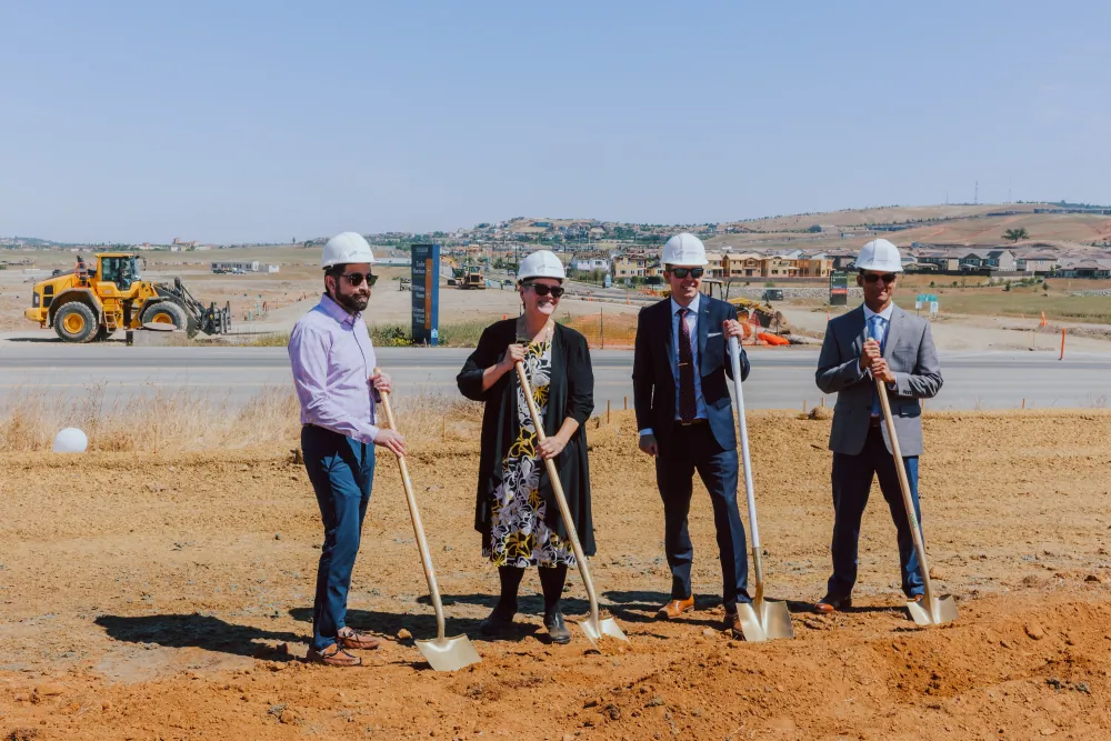 Four officials in hard hats hold ceremonial shovels at the Capital SouthEast Connector groundbreaking, with a loader and graded roadway behind them