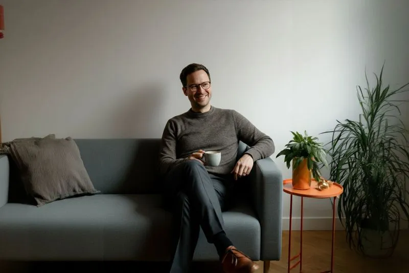 Man wearing glasses sitting on a gray sofa, holding a coffee cup, smiling.
