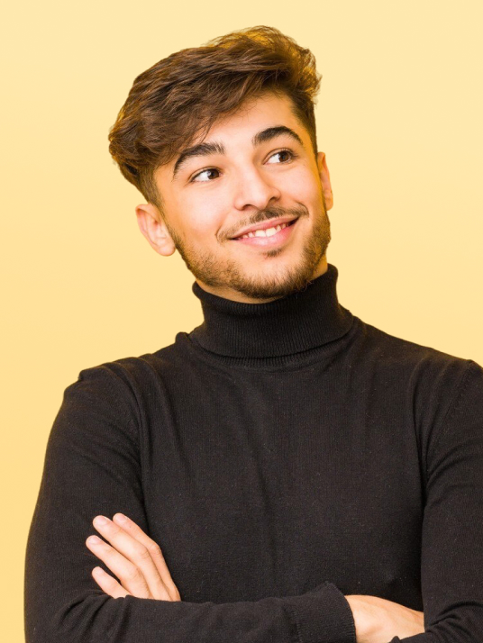 Young man with brown hair and beard wearing a black turtleneck sweater, smiling and looking to the side with arms crossed against a yellow background.