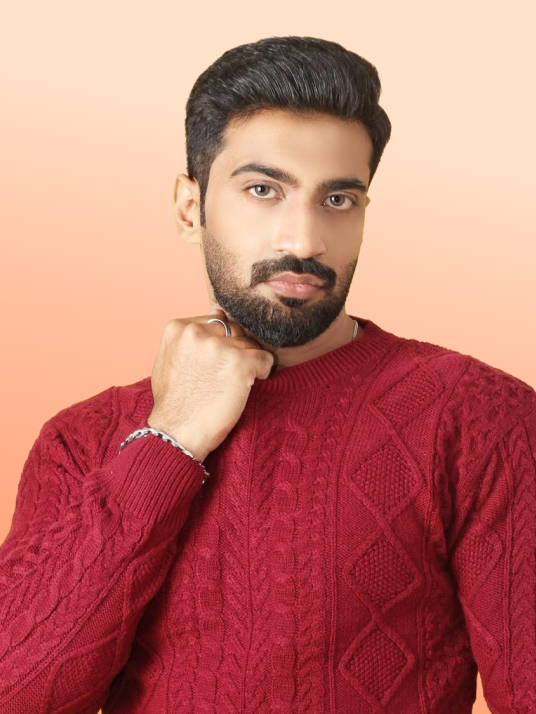 Man with dark hair and beard wearing a red cable-knit sweater, posing with his fist near his chin against a peach-colored background.