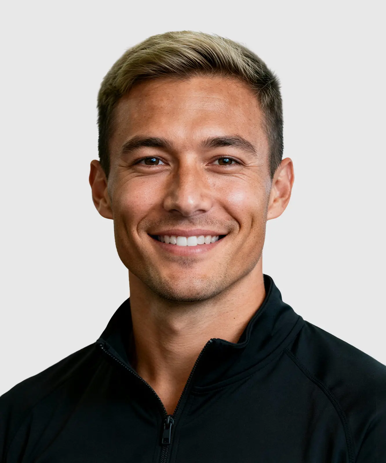 Smiling fit young man with short blond hair in a black zip-up jacket in a gym setting.