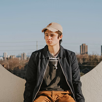 Young man in a beige cap and black jacket sitting outdoors with city buildings in the background.