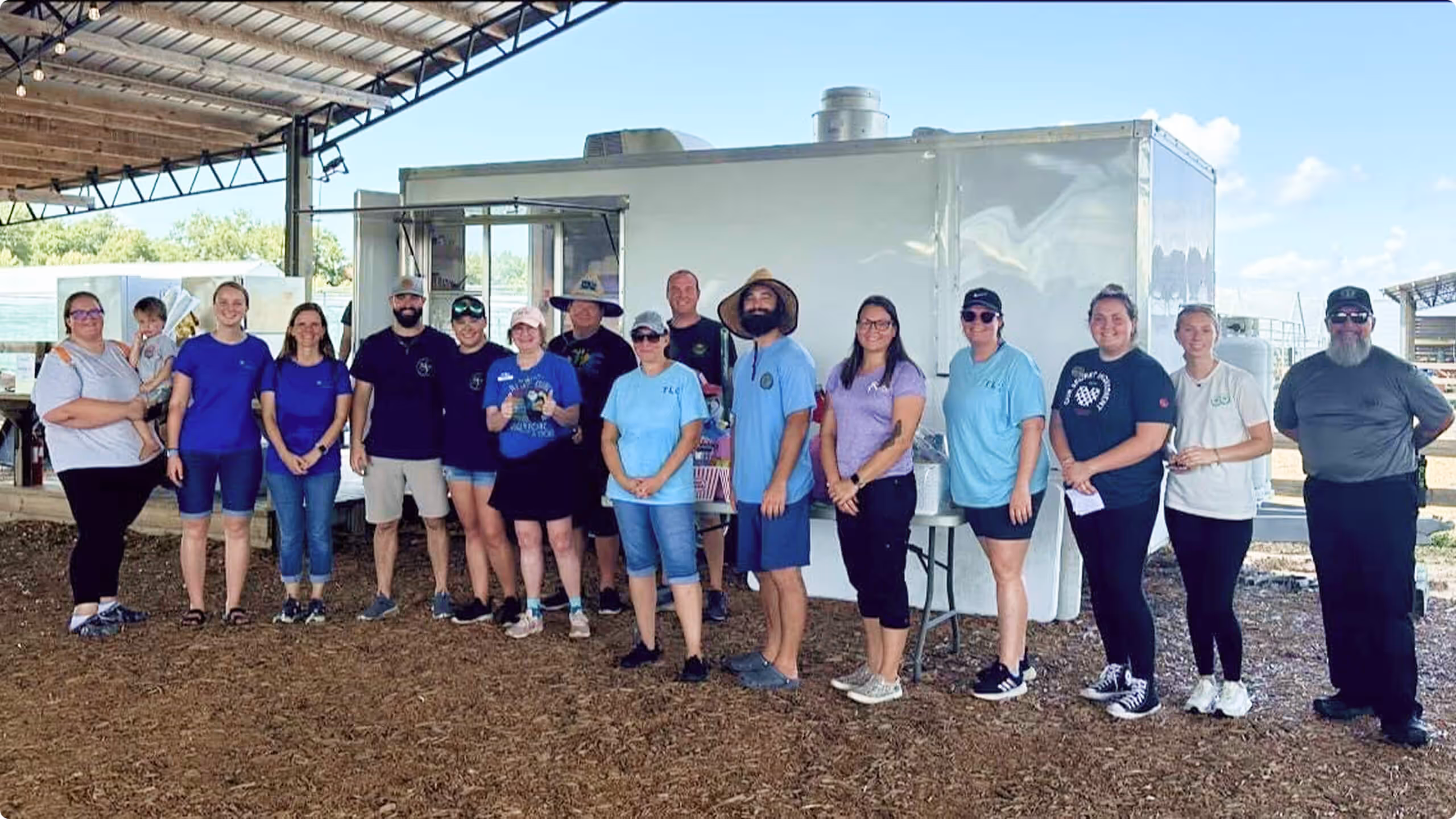 Group of sixteen people standing outdoors under a roof, smiling in front of a white trailer.