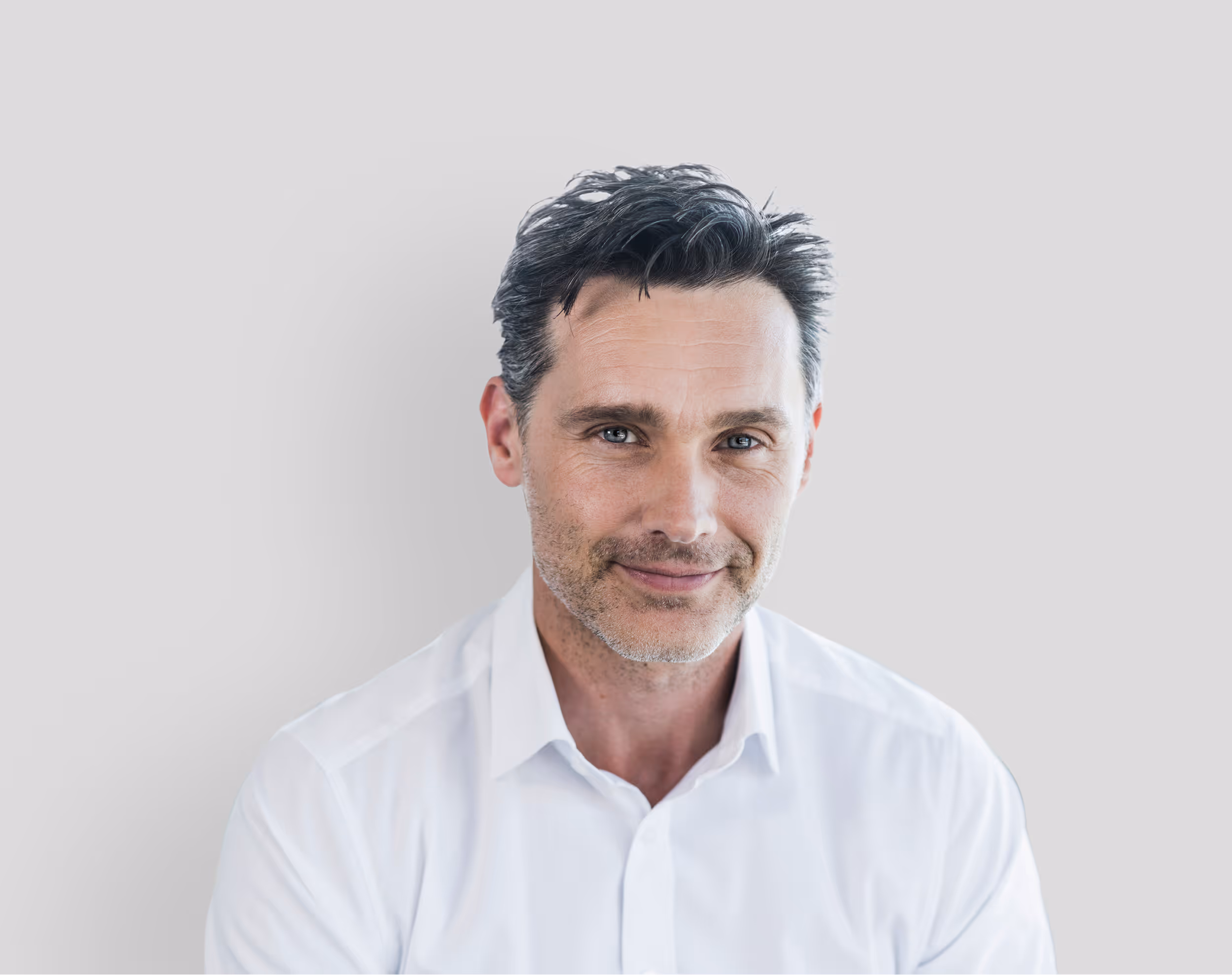 Smiling middle-aged man with dark hair wearing a white button-up shirt against a plain light gray background.