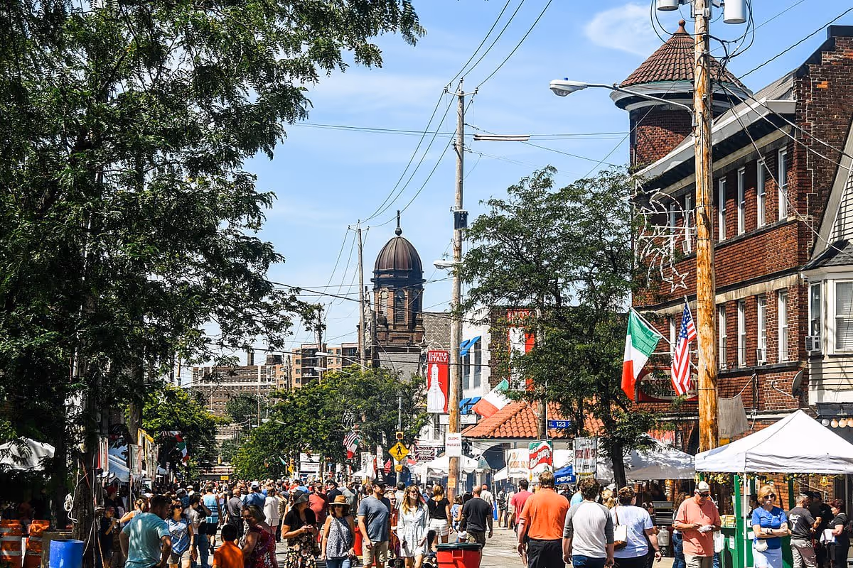 Crowded street fair in a neighborhood with Italian and American flags, tents, and a historic brick building with a domed tower.