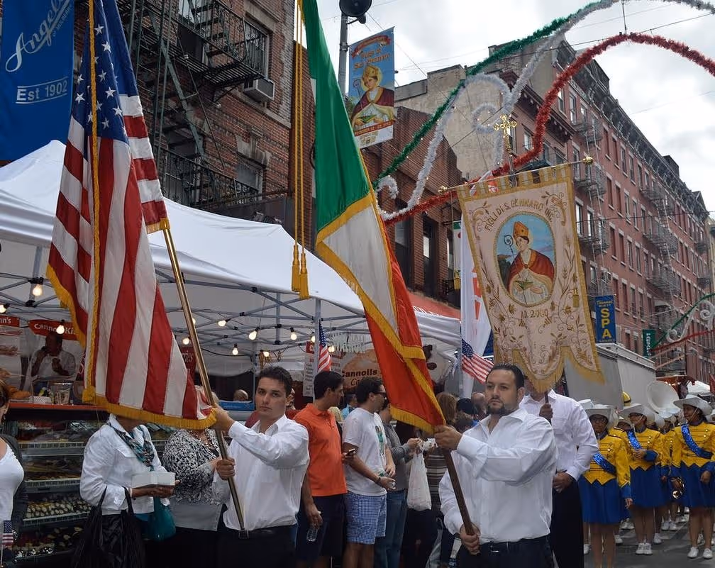 Two men in white shirts carry American and Italian flags during a street parade with people and musicians in yellow and blue uniforms.