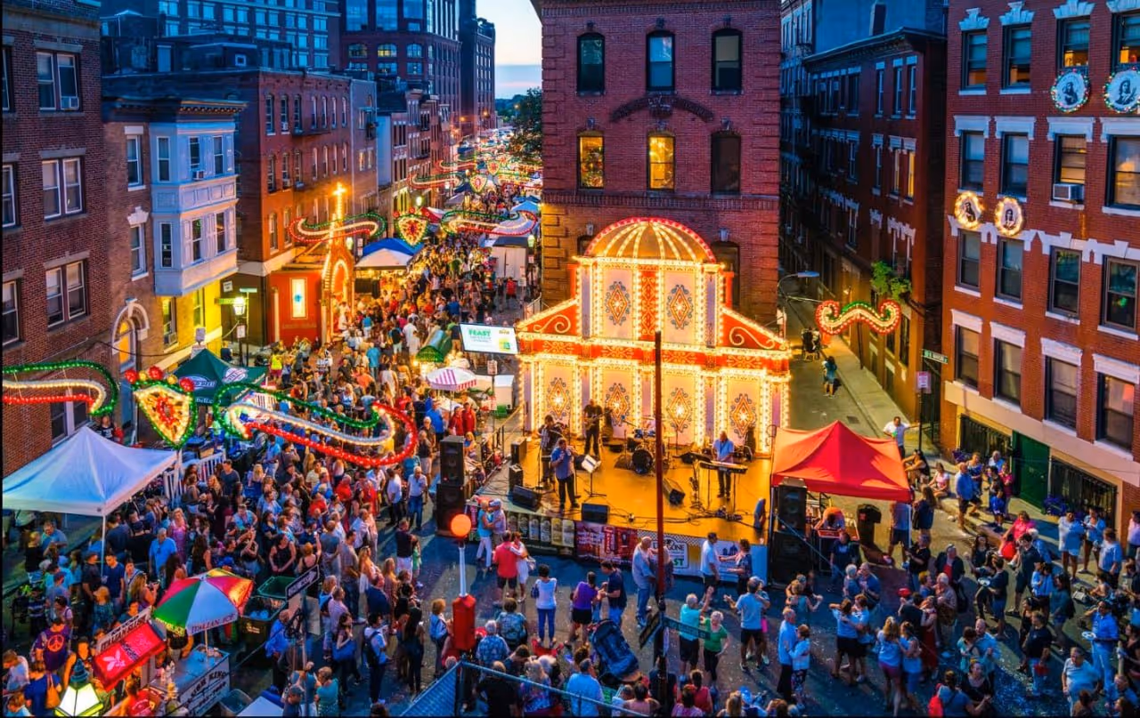 Crowded street festival with people gathered around a brightly lit stage playing live music between brick buildings at dusk.