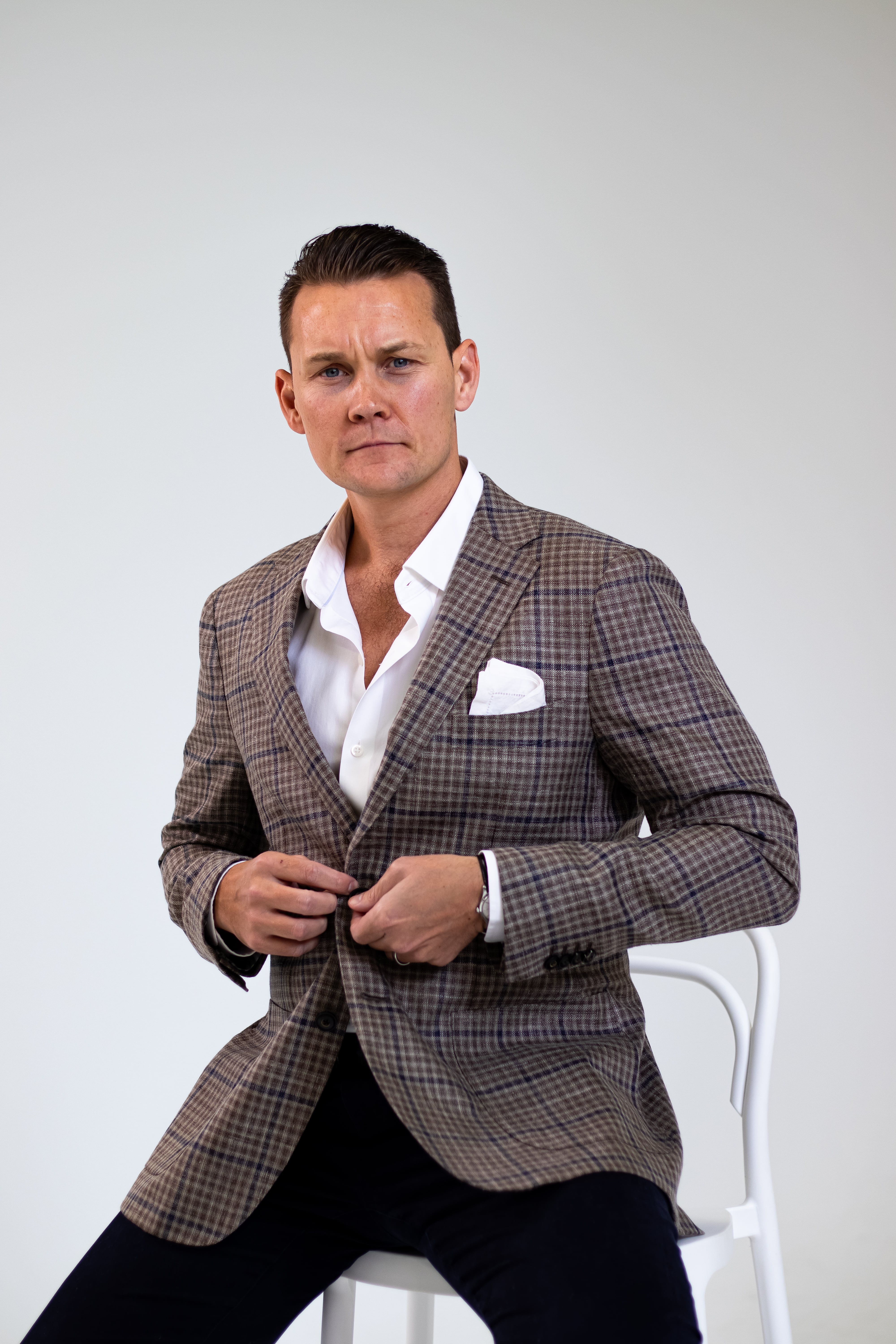 An image of a man sitting on a white chair wearing a pleaded blazer looking directly at the camera