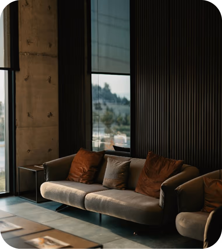 Modern living room with a gray sofa and two brown pillows, a gray cushion in the center, and a matching armchair against a dark ribbed wall near large windows.