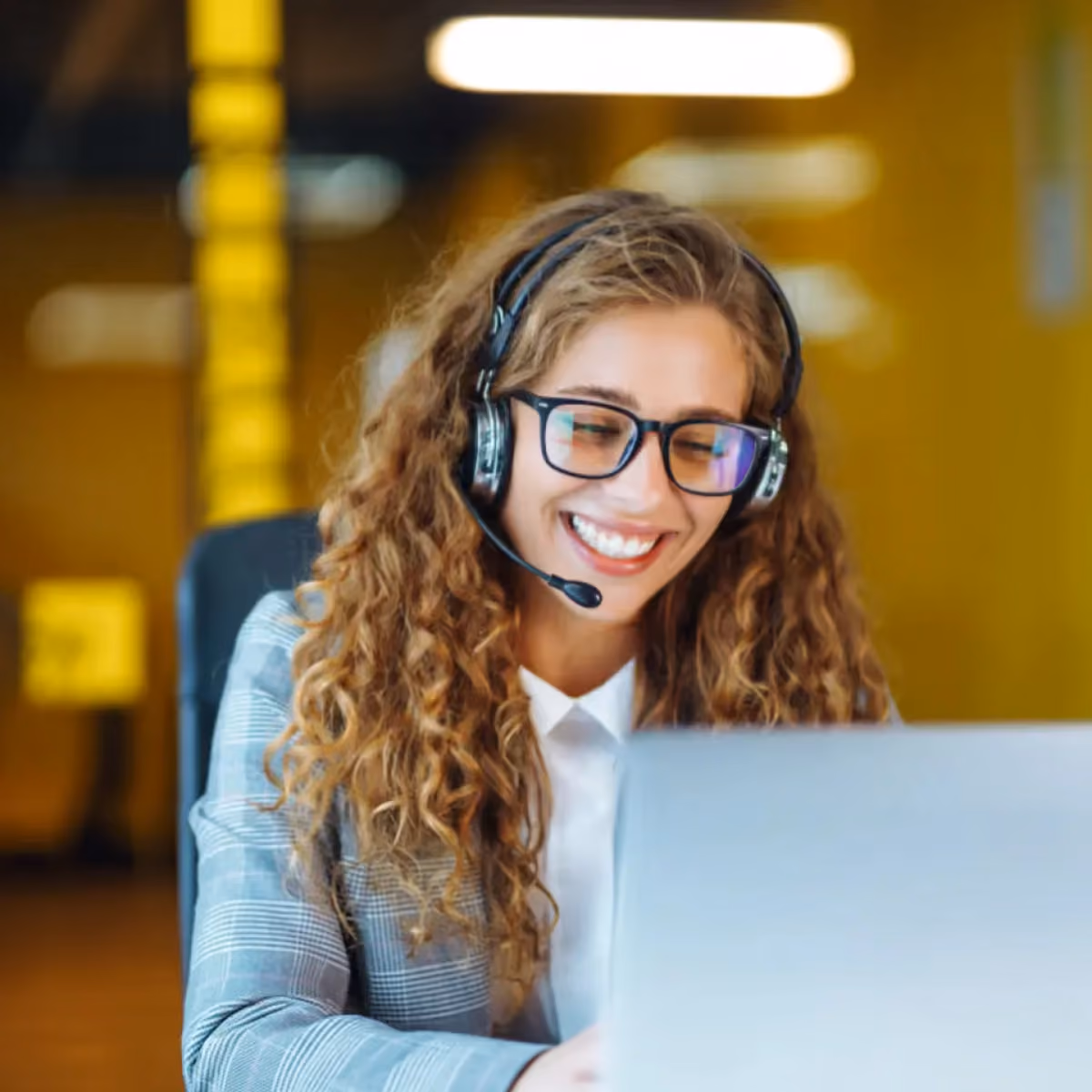 Lächelnde Frau mit lockigem Haar, Headset und Brille arbeitet an einem Laptop in einem gelben Büro.