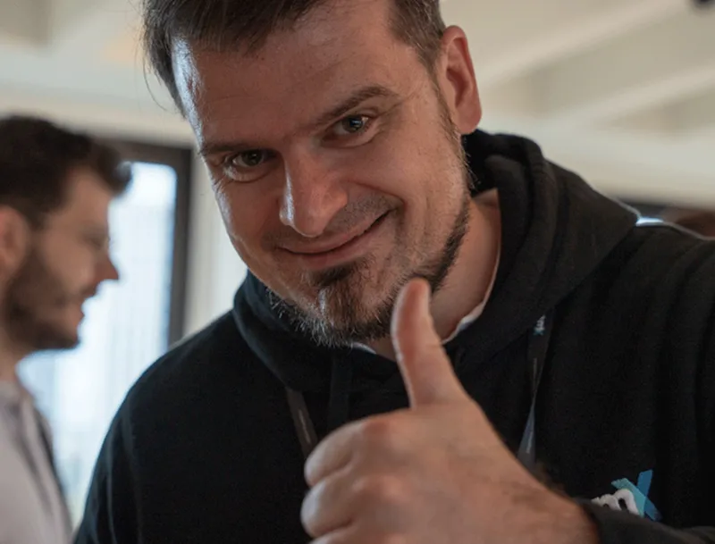 Man in black hoodie smiling and giving thumbs-up with blurred man in the background.