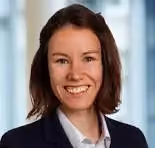 Lächelnde Frau mit schulterlangem braunem Haar, dunklem Blazer und weißem Hemd.