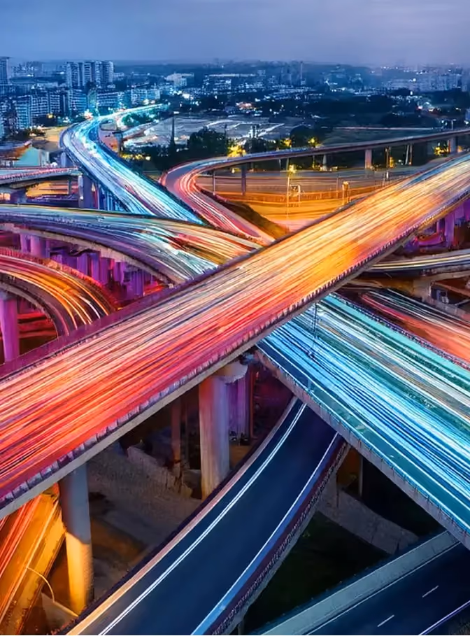 Erhöhtes Autobahnkreuz bei Nacht mit Lichtspuren vorbeifahrender Fahrzeuge.