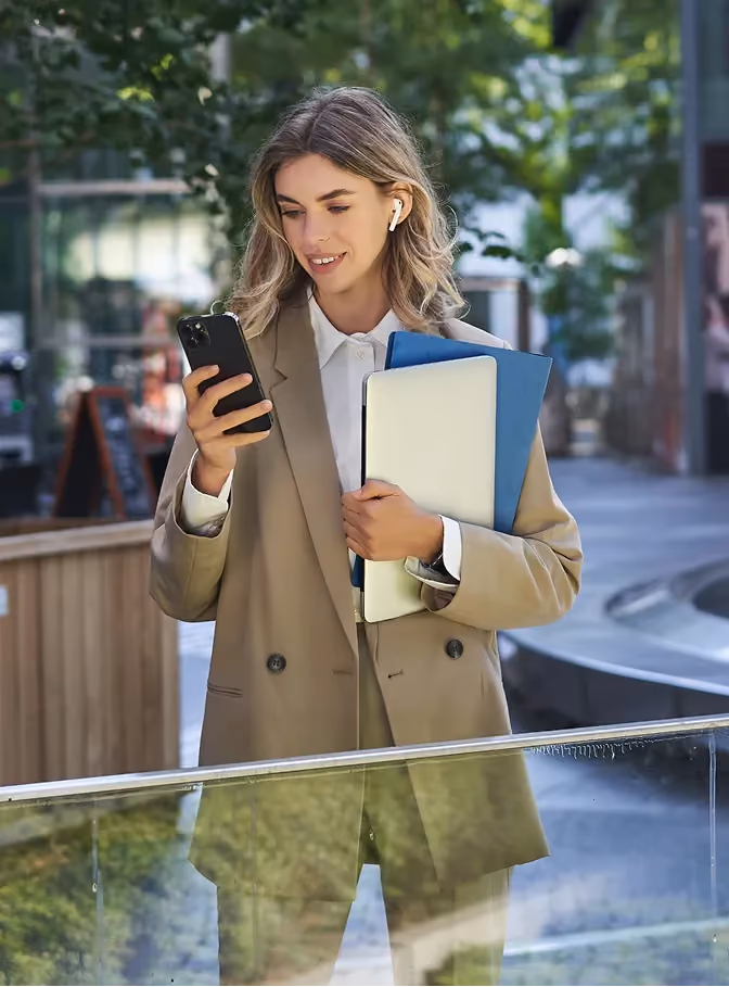 Lächelnde Frau im beigen Anzug nutzt ihr Smartphone und hält Laptop und Mappe im Freien.