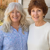 Two smiling middle-aged women with their arms around each other standing outdoors in front of a porch.