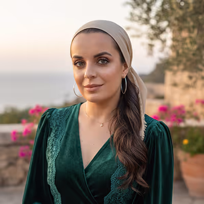 Woman with long wavy brown hair wearing a beige headscarf and green velvet dress, standing outdoors with pink flowers and stone walls in the background.