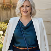 Smiling blonde woman wearing a white blazer over a dark green blouse standing outdoors in front of a beige wall.