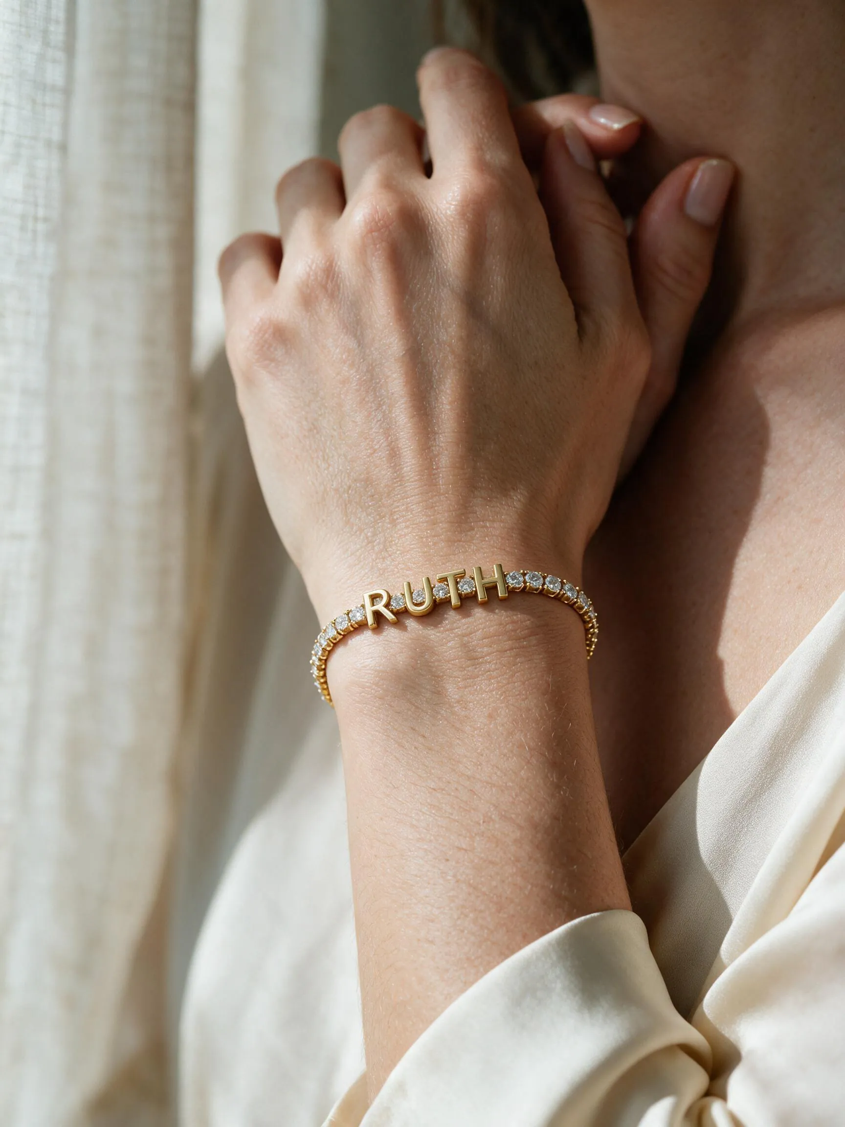 The RUTH bracelet - a gold tennis bracelet with diamond links and gold letters spelling Ruth, worn on a wrist held to the collarbone