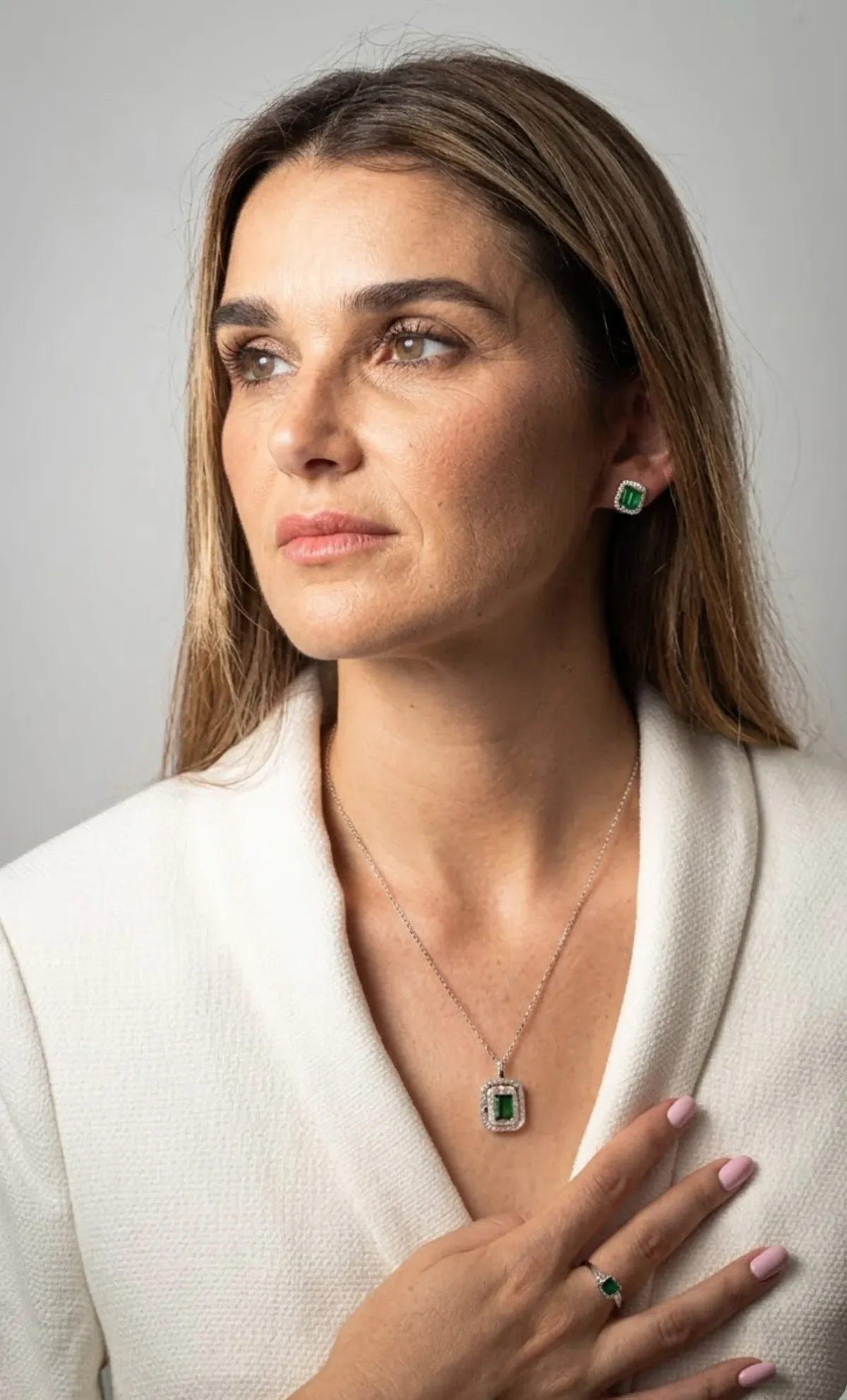 Woman in white textured blazer wearing matching emerald green and diamond earrings, necklace, and ring.