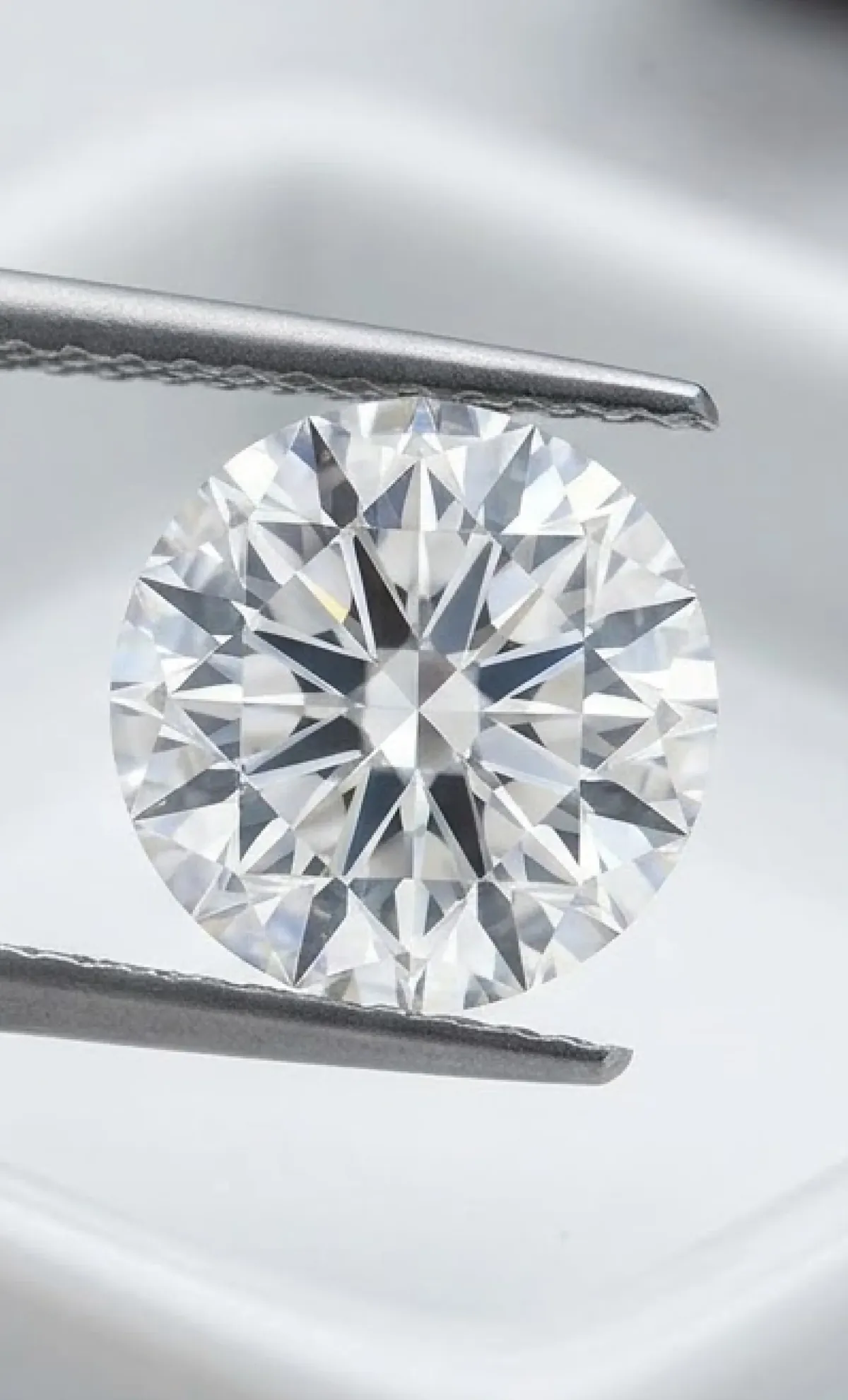 Close-up of a round brilliant-cut diamond held by metal tweezers against a soft gray background.