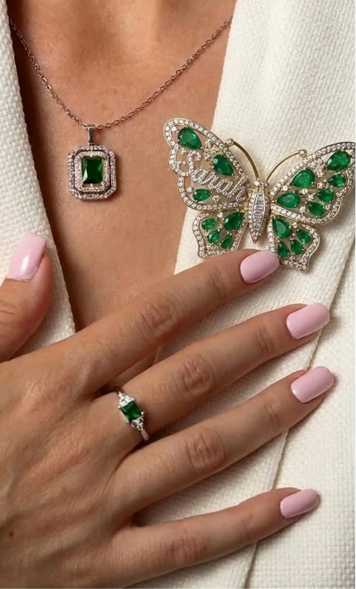 Hand with pink nails wearing a silver ring with a green gemstone, near a white jacket adorned with a butterfly-shaped brooch and a matching green gemstone pendant necklace.