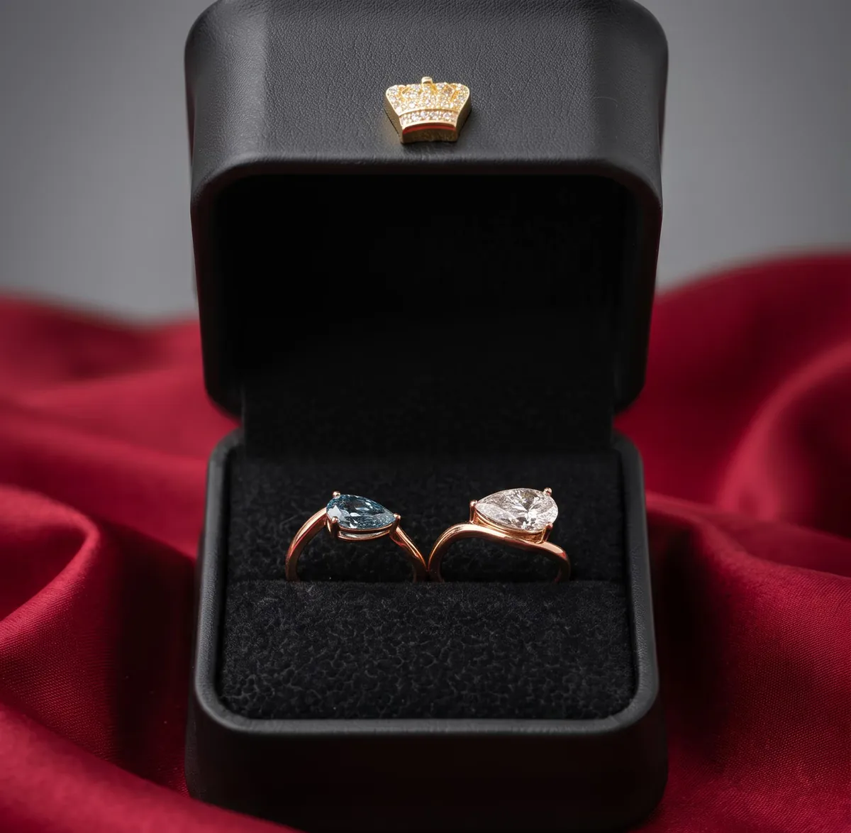 Two rose gold rings with pear-shaped gemstones, one blue and one clear, in an open black ring box with a gold crown emblem on top, against a red fabric background.