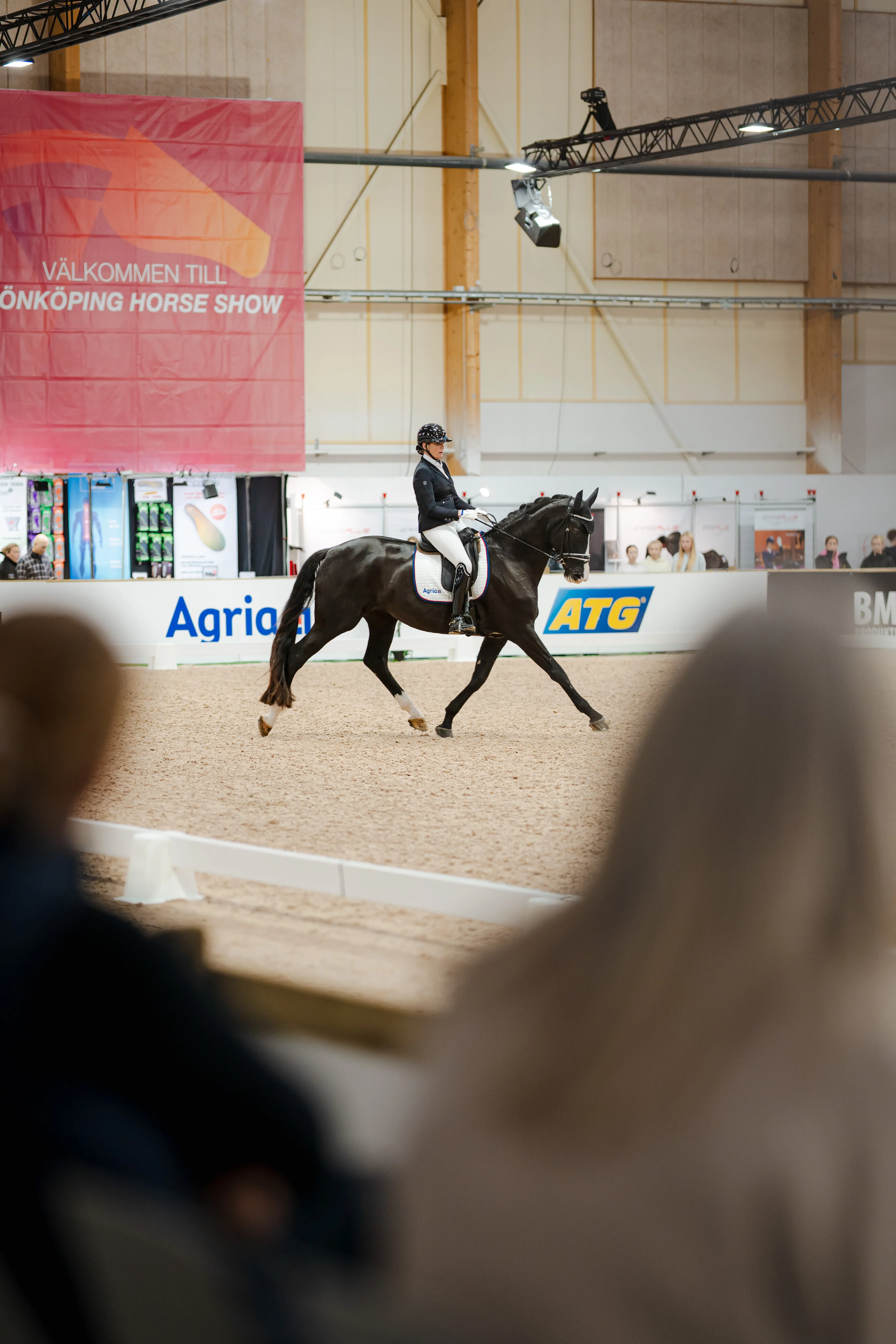 Rider in formal attire performing dressage on a black horse inside an indoor arena with spectators.