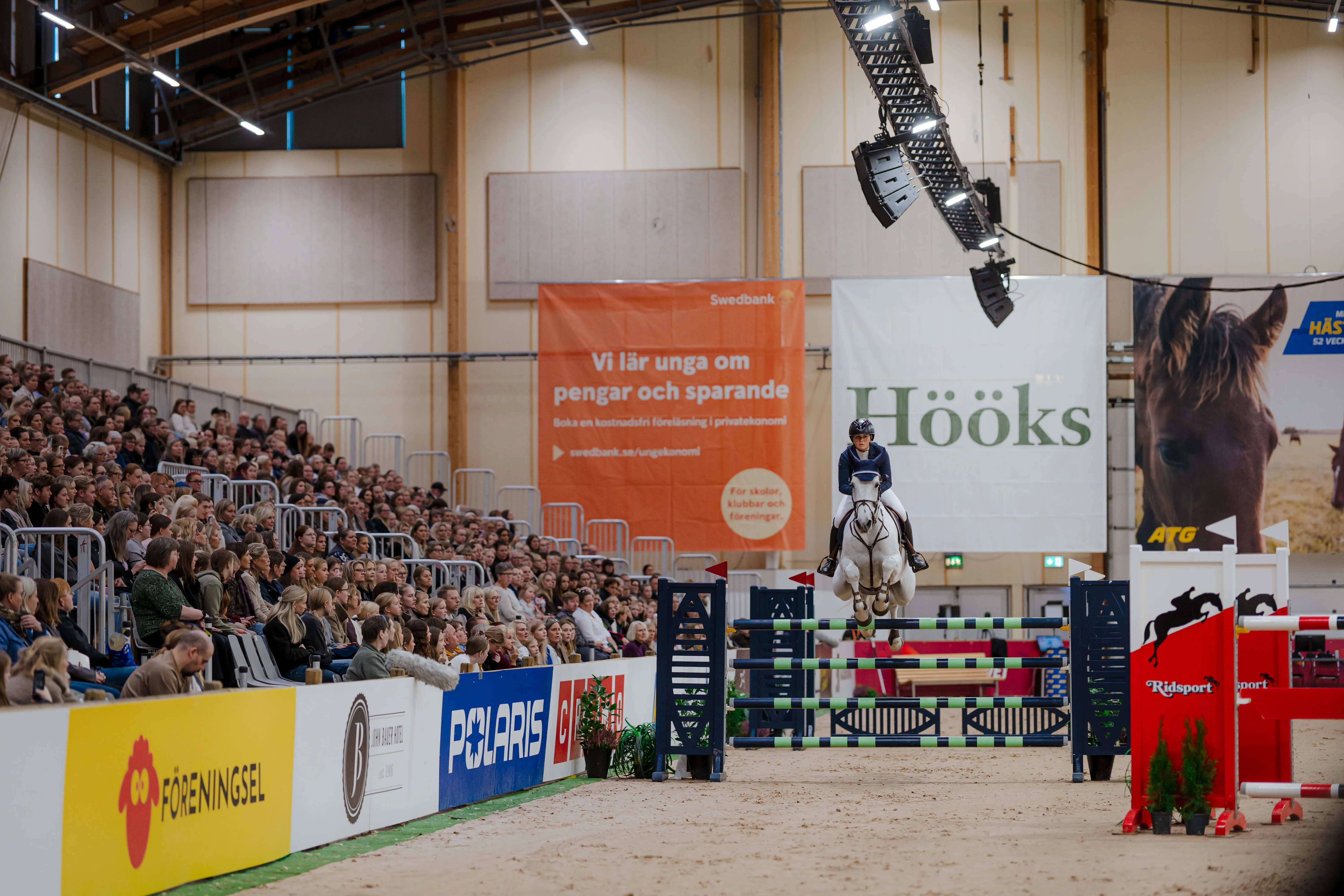 Equestrian rider jumping a white horse over an obstacle indoors with a large audience watching.