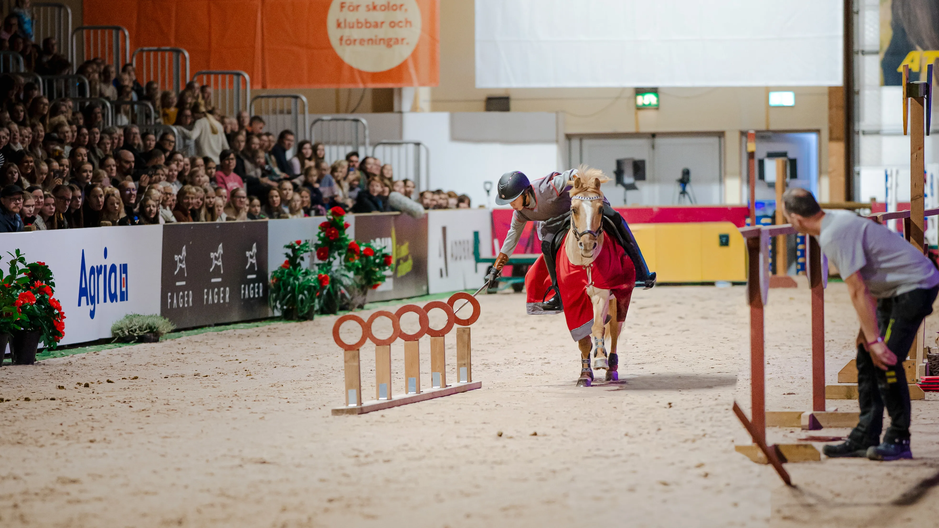 Person on horse in red coat leans to hit ring in equestrian riding competition with audience watching.