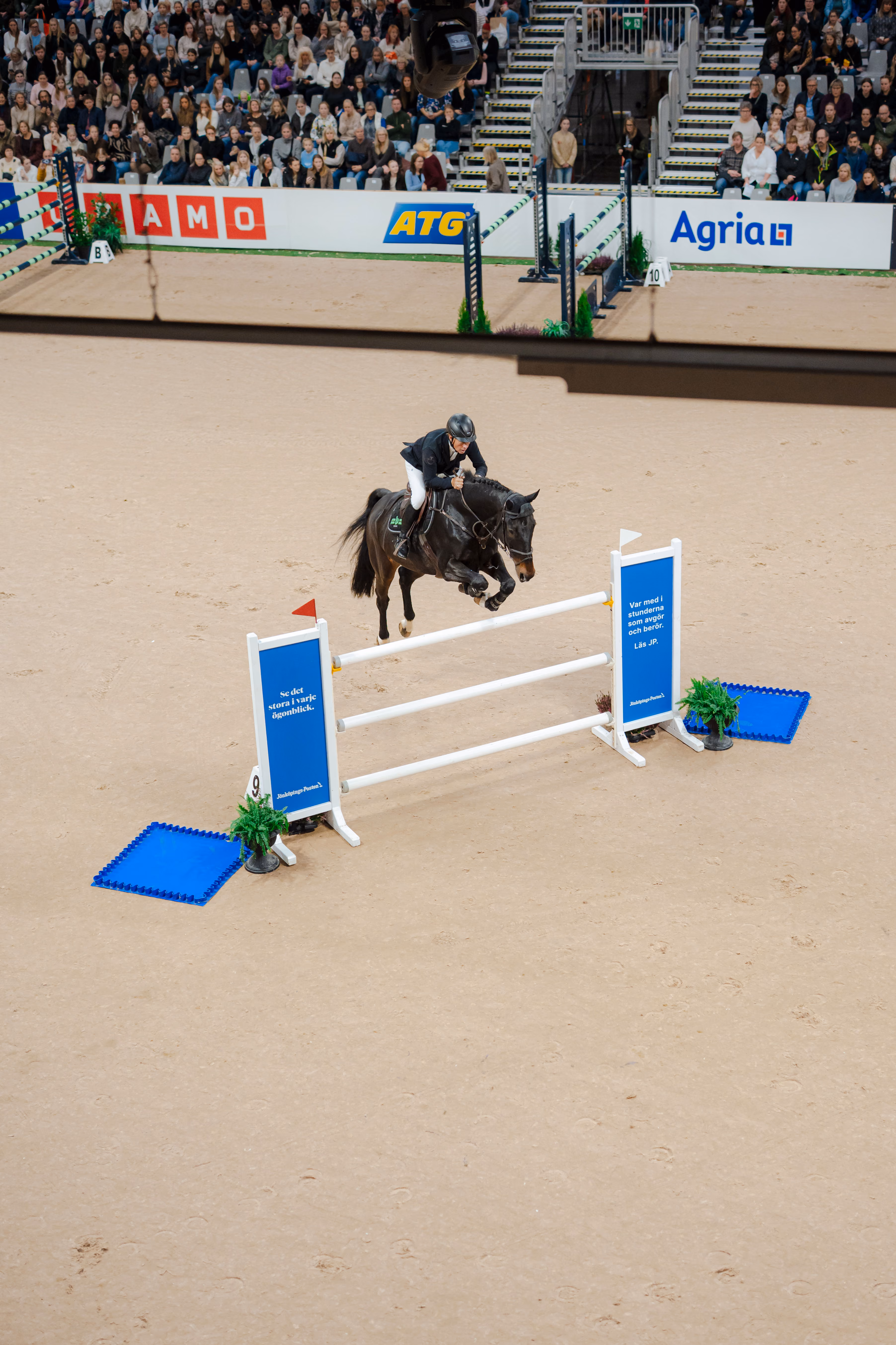 Horse and rider in mid-air jumping over an obstacle in an indoor equestrian event with audience.